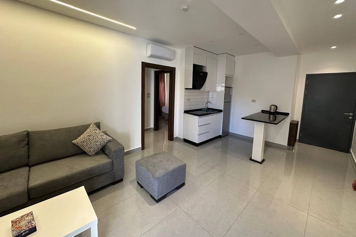 Een moderne woonkamer met een bank, een poef en een klein tafeltje. In de achtergrond is een keuken met witte kasten en een zwart aanrecht zichtbaar.