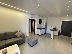 A modern living room with a sofa, ottoman, and a small table. A kitchen with white cabinets and a black countertop is visible in the background.