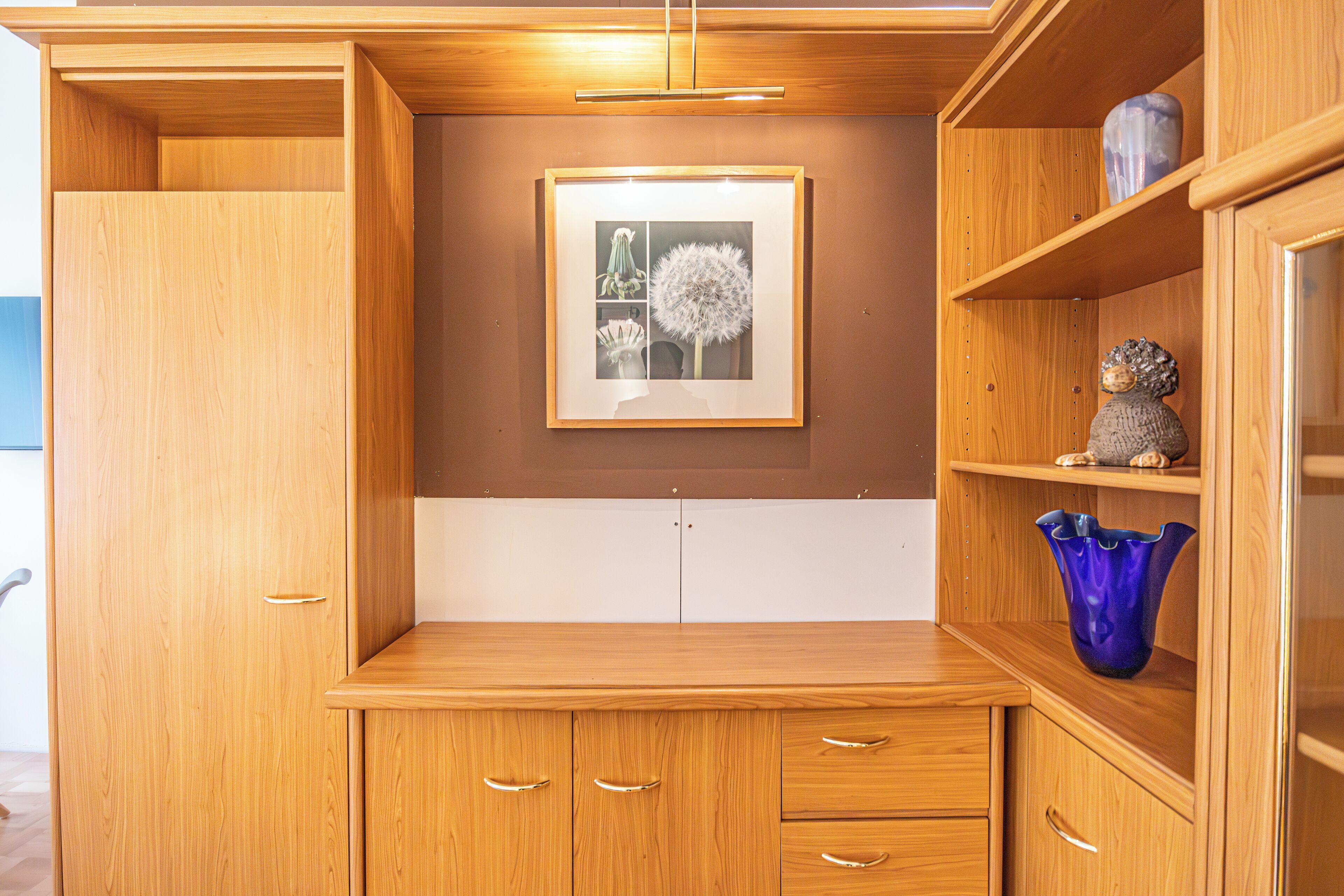 A wooden cabinet with multiple drawers and shelves, a framed picture on the wall, and a blue vase on the top shelf.