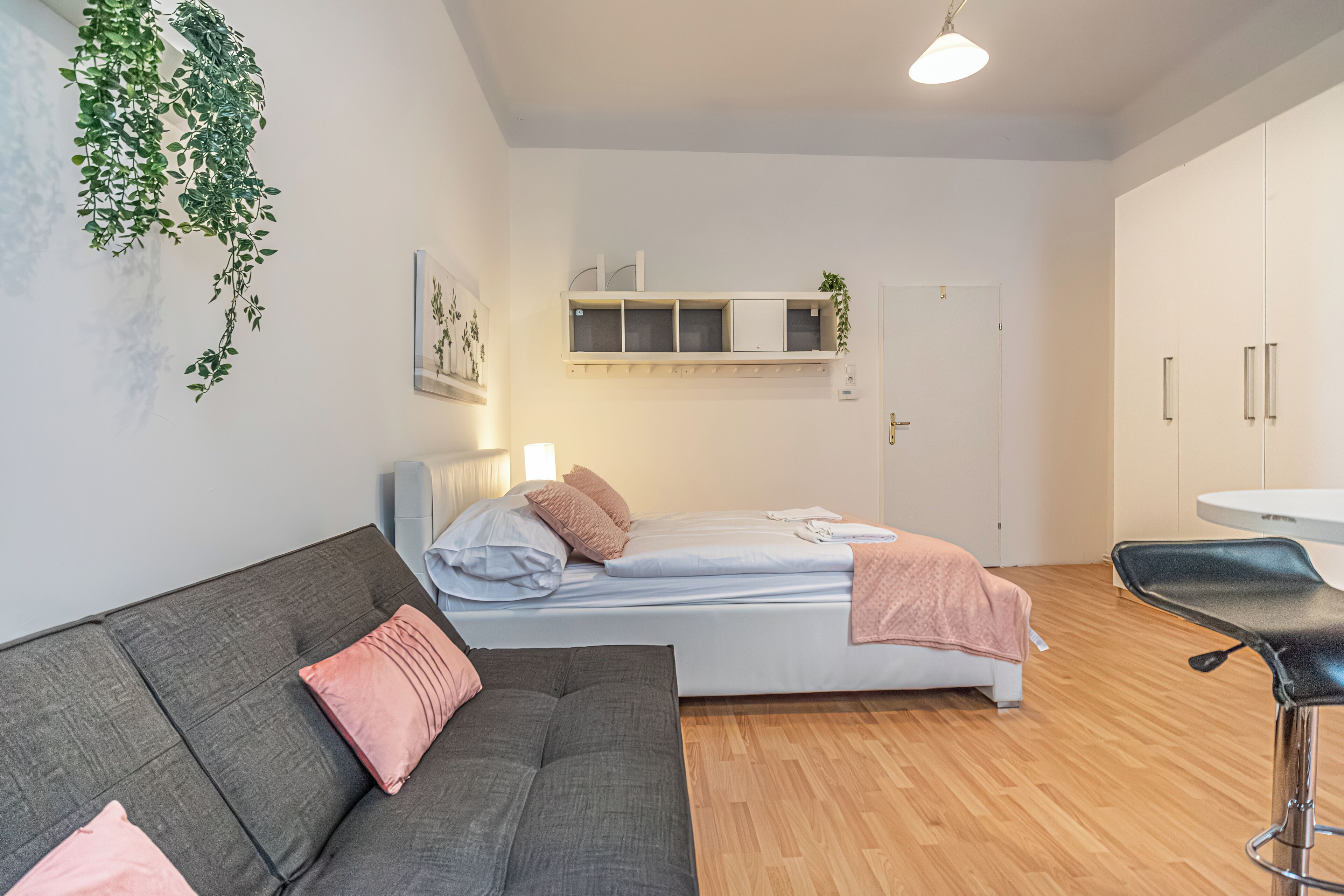 A modern bedroom with a bed, a grey sofa, a small desk, and a hanging plant.