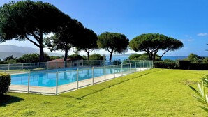 Une piscine entourée d’une clôture en verre, bordée d’une pelouse et d’arbres bien entretenus, offrant une vue dégagée sur la mer et les montagnes à l’arrière-plan.