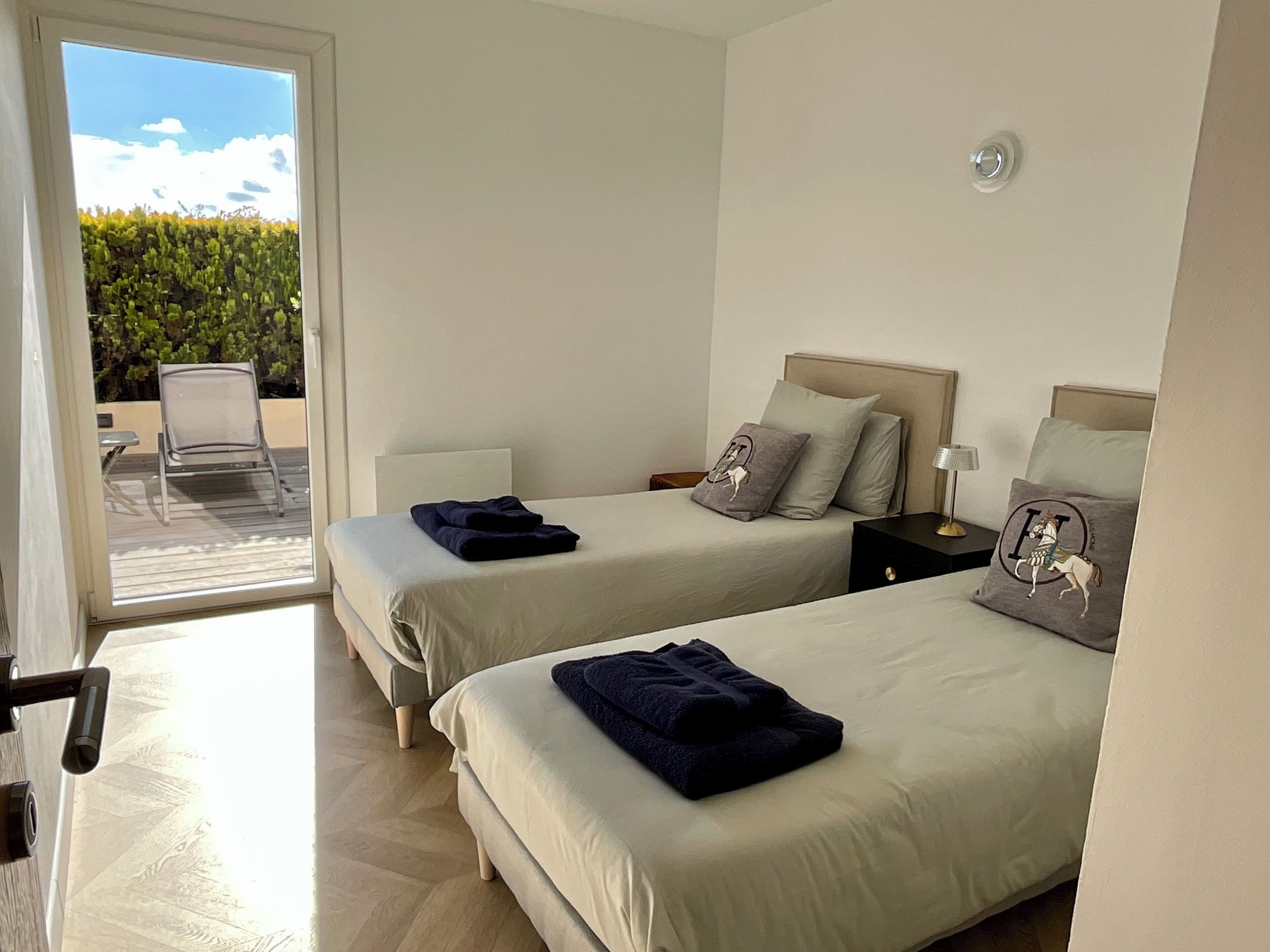 A hotel room with two beds, a small table, a chair, and a view of greenery outside.