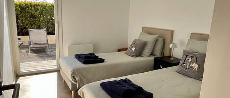 A hotel room with two beds, a small table, a chair, and a view of greenery outside.