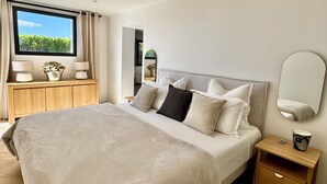 Une chambre moderne avec un grand lit, des tables de chevet, une commode, un miroir et une fenêtre donnant sur un paysage verdoyant.