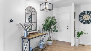 Un couloir doté d’un plancher en bois, d’une console en métal agrémentée d’objets décoratifs, d’un grand miroir, d’une plante en pot et d’une horloge murale.