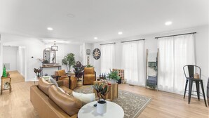 Un salon moderne avec un canapé en cuir marron, une table basse ronde et un grand miroir. On y trouve une table d’appoint en bois sur laquelle sont posés une plante et un petit sapin de Noël. Les murs sont blancs, le sol est en bois et une grande fenêtre est agrémentée de rideaux blancs.