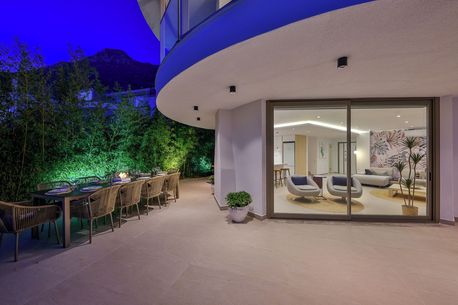 A modern outdoor dining area with wicker chairs and a table, surrounded by lush greenery and a mountain backdrop.