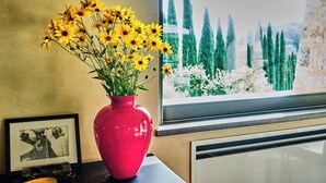 A window with a view of tall cypress trees and a cross, a pink vase with yellow flowers, and a framed picture on a table. - Godiluva, wine villa (Toscana)