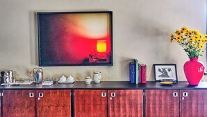 Un comptoir de cuisine avec une machine à café, une théière et des tasses. Un tableau encadré est accroché au-dessus du comptoir, et un vase rempli de fleurs jaunes se trouve à droite.