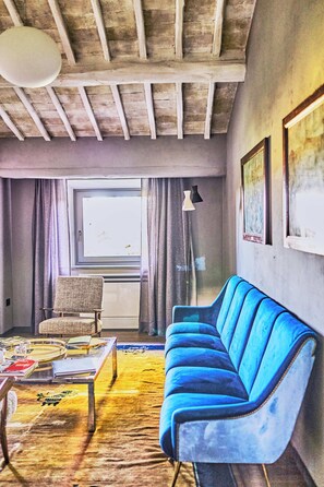 Une pièce avec un canapé bleu, un fauteuil, une table avec des livres et un verre, une fenêtre avec des rideaux et une lampe.