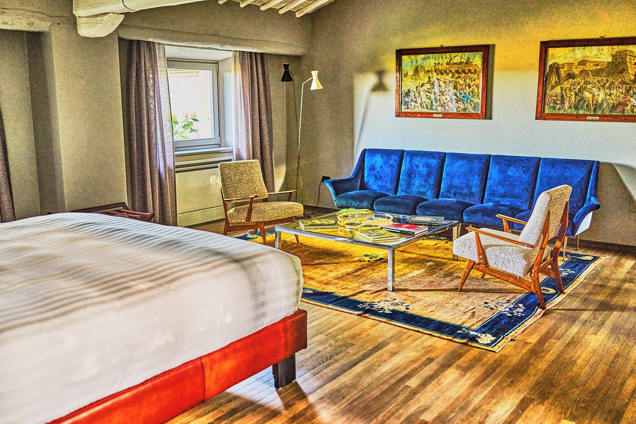 Ein Hotelzimmer mit einem Bett, einem blauen Sofa, zwei Sesseln, einem Couchtisch und einem großen Fenster mit Vorhängen.