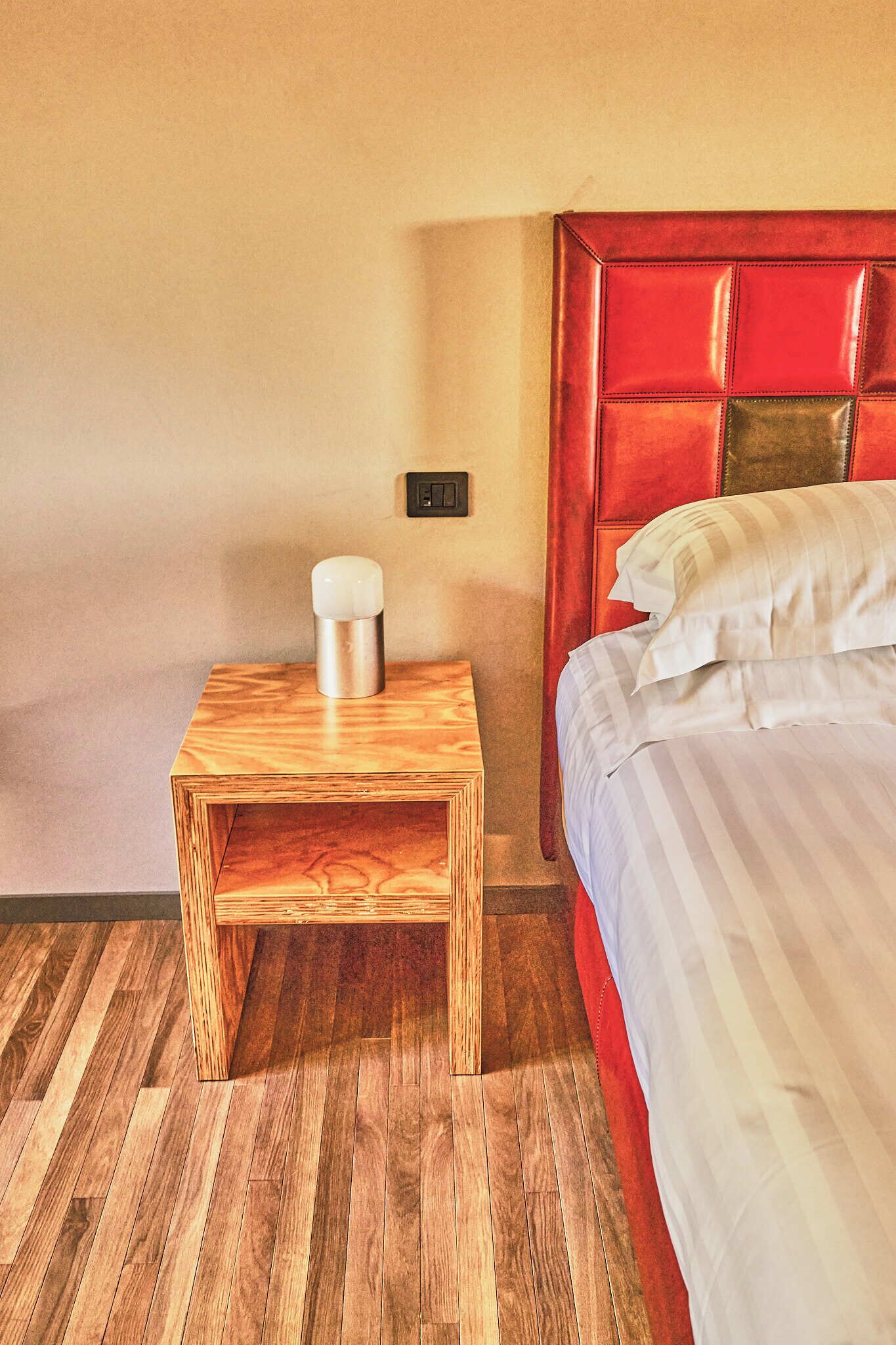 Ein Hotelzimmer mit einem hölzernen Nachttisch, einem Bett mit weiß gestreifter Bettwäsche und einem roten Kopfteil.