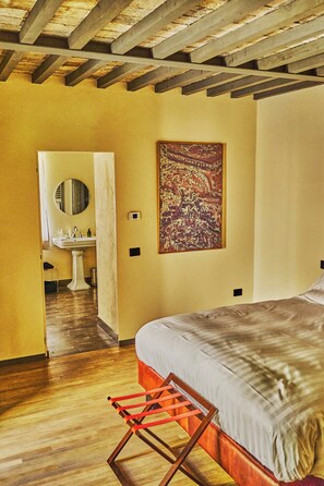 Une chambre à coucher avec un lit, un plancher en bois, une salle de bain avec un lavabo et un miroir, et un tableau accroché au mur.