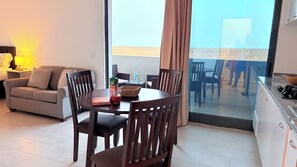 A modern living room with a sofa, a dining table with chairs, and a view of the beach through large windows.