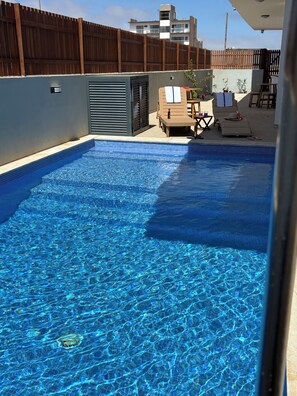 A clear blue pool with lounge chairs and a table, surrounded by a wooden fence and buildings in the background. - JoLi Beach House - Spacious Second Floor T0 Suite With Beatiful View (Santa Maria)