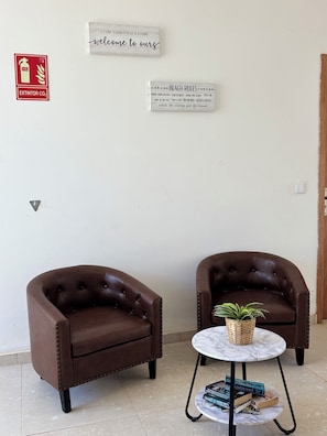 Deux fauteuils en cuir marron, une petite table ronde avec une plante et des livres, et deux panneaux accrochés au mur : l’un pour accueillir les visiteurs et l’autre indiquant les règles à respecter sur la plage.