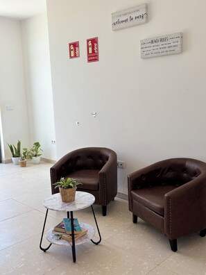 A room with two brown leather armchairs, a small round table with a plant, and a marble floor. There are two red signs on the wall, one with a fire extinguisher symbol and another with a beach rules message. - JoLi Beach House - Second Floor T1 Beautiful and Authentic Island Living (Santa Maria)