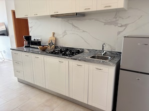 Una cocina moderna con armarios blancos, salpicadero de mármol, estufa a gas, pileta y refrigerador.