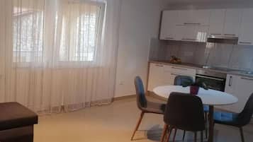 Ett kompakt bostadsutrymme med ett litet kök, en matplats med ett runt bord och stolar samt ett stort fönster med tunna, genomskinliga gardiner.
