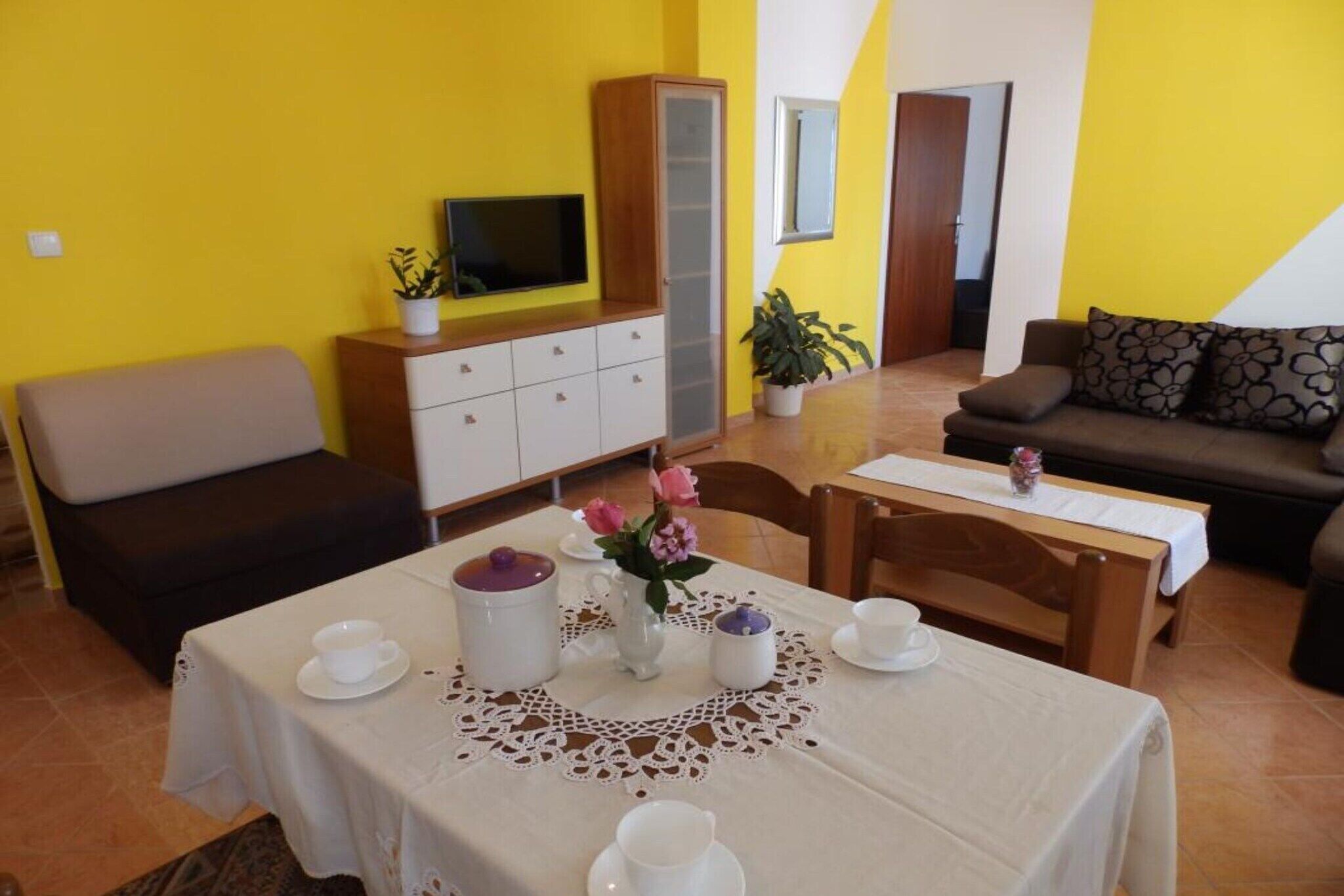 A dining area with a table set for two, a cabinet with a television, and a sofa in a room with yellow walls.