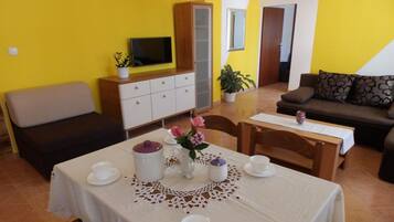 A dining area with a table set for two, a cabinet with a television, and a sofa in a room with yellow walls.