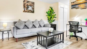 A modern living room with a grey sofa, patterned cushions, a black coffee table, a small side table with a lamp, a green plant, and a desk with a black office chair. - TruStay in Central CT (Manchester)