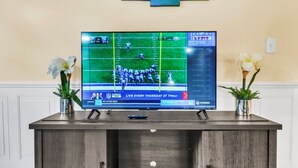 A television displaying a football game, placed on a wooden stand with two potted plants on either side. - TruStay in Central CT (Manchester)