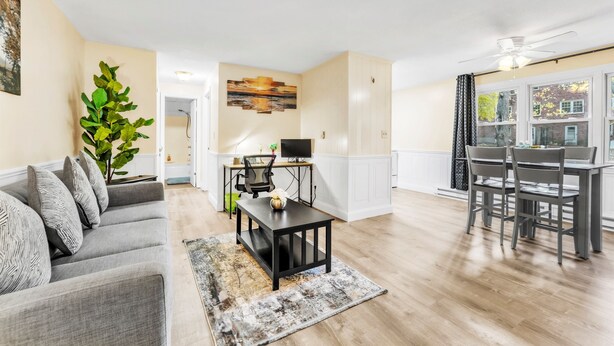 A modern living room with a gray sofa, a coffee table, and a dining area with a table and chairs. There is a TV and a plant in the room. - TruStay in Central CT (Manchester)