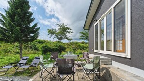 Eine Terrasse im Freien mit Liegestühlen und einem Tisch, umgeben von Grünpflanzen und einem Haus mit großen Fenstern.