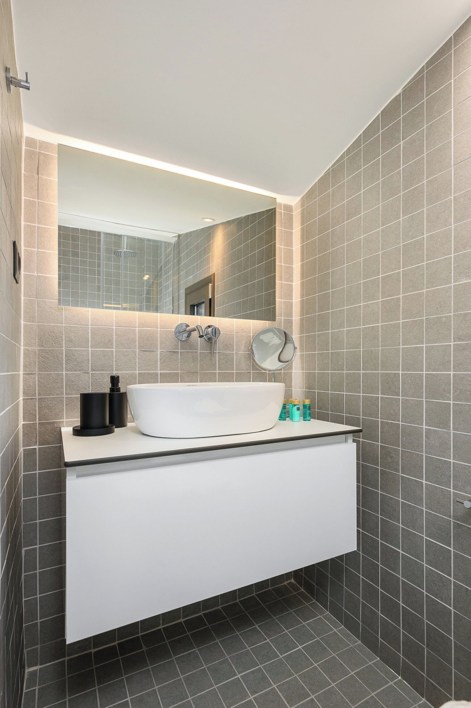A modern bathroom with a white sink, a round mirror, and a large mirror above the sink. The walls are tiled in a light gray color, and there is a small shelf with toiletries.
