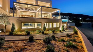 A modern two-story house with a covered patio, large windows, and a landscaped garden. - Majo Luxury 11BDR Villa with Sea View 2Heated Pools, Gym Sauna (Heraklion)