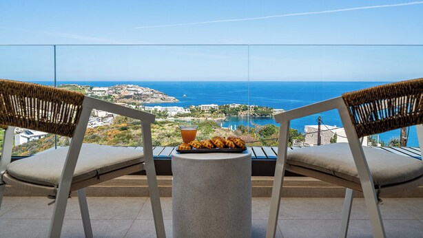A balcony with two wicker chairs, a small table, and a tray of food and drink. The view includes a coastal town and the sea. - Majo Luxury 11BDR Villa with Sea View 2Heated Pools, Gym Sauna (Heraklion)