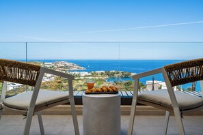A balcony with two wicker chairs, a small table, and a tray of food and drink. The view includes a coastal town and the sea. - Majo Luxury 11BDR Villa with Sea View 2Heated Pools, Gym Sauna (Heraklion)