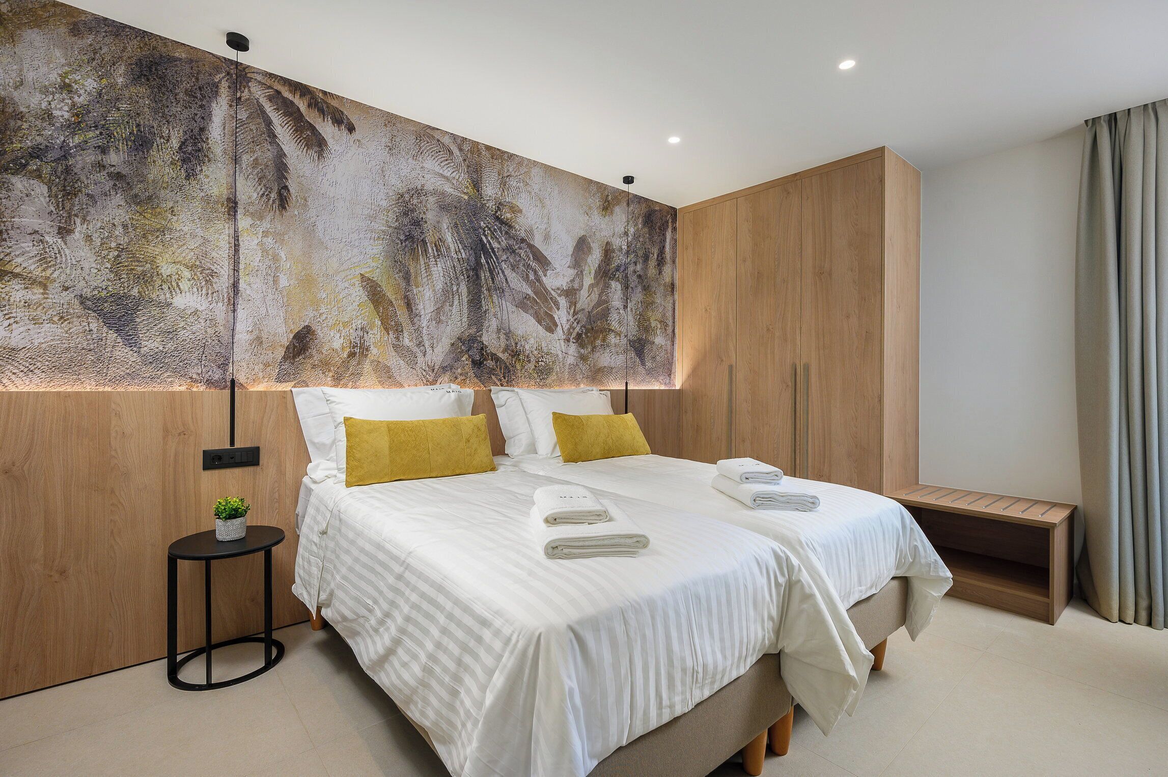 A modern hotel room with a large abstract mural on the wall, two beds with white bedding and yellow pillows, a small round table with a plant, and a wooden bench.