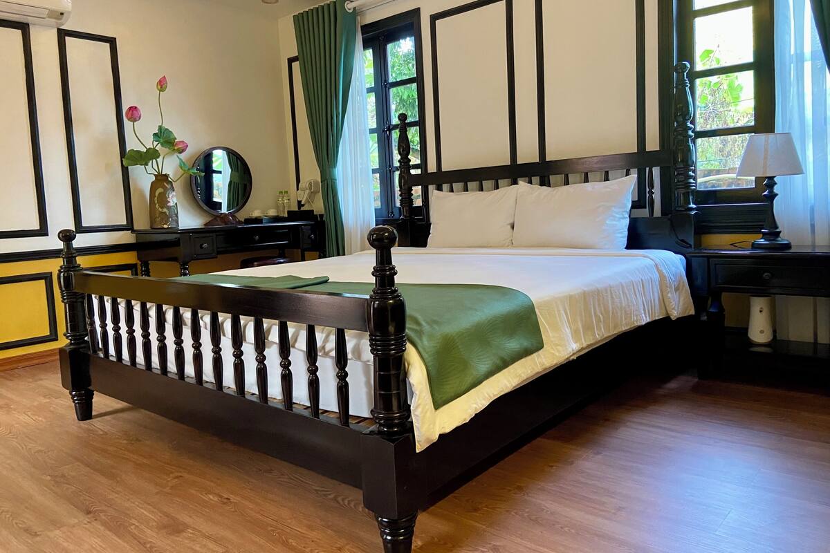Ein traditionelles Himmelbett mit grüner Tagesdecke, ein Nachttisch aus Holz mit Lampe, eine Kommode mit Spiegel und ein Fenster mit Vorhängen.