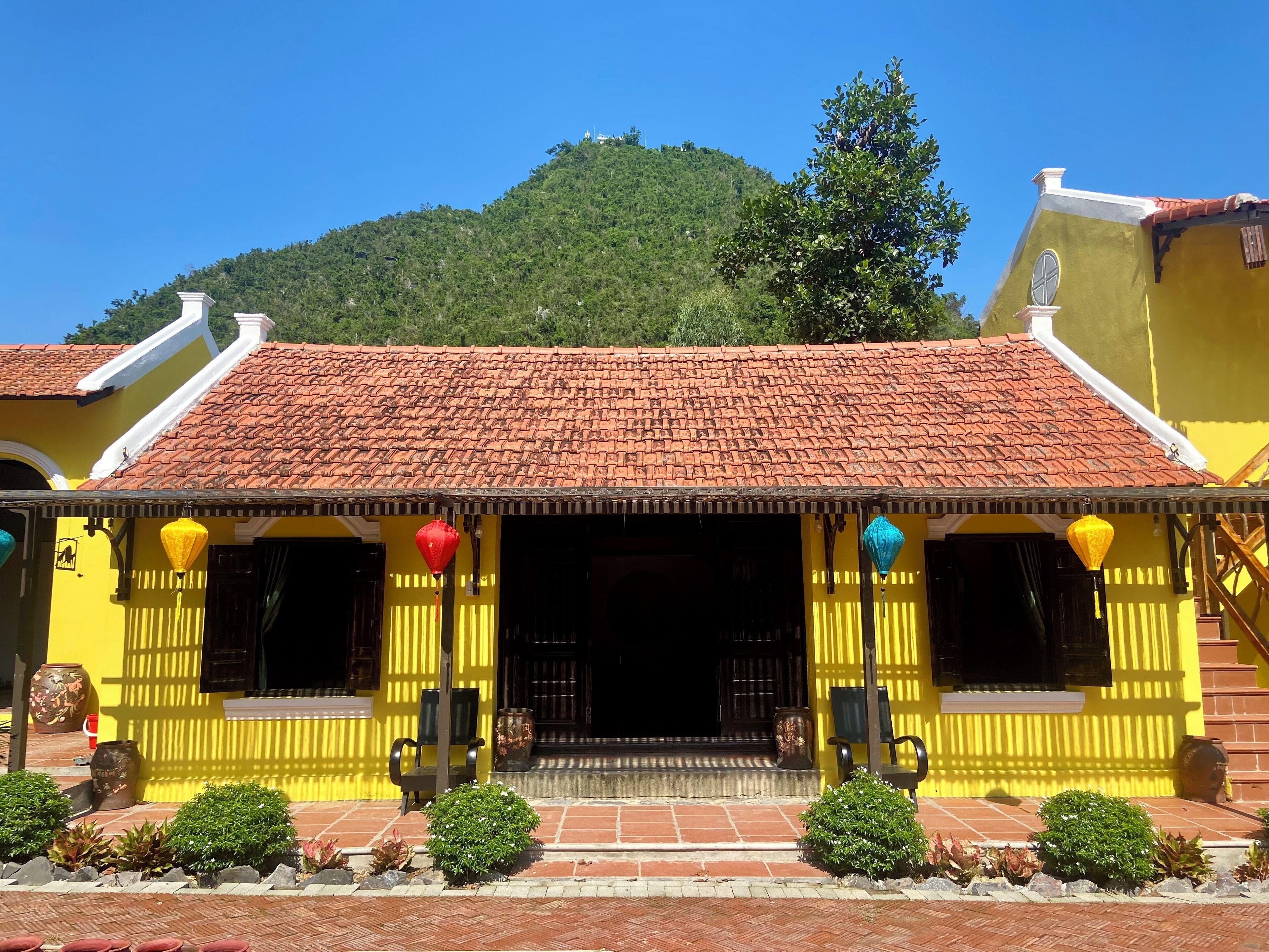 A yellow building with a red-tiled roof, a staircase, and a view of a green hill in the background.