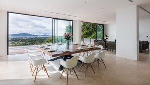 A modern dining area with a large table set for six, large windows offering a scenic view, and a balcony with wicker furniture. - Stunning Views -  Villa Maan Ingfah VMI1 (Chalong)