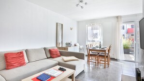 Ein modernes Wohnzimmer mit einer Couch, einem Esstisch und Stühlen. Ein Fernseher ist an der Wand befestigt und ein Fenster sorgt für natürliches Licht.