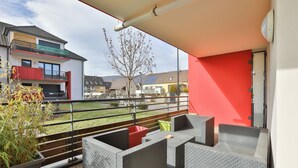 Ein Balkon mit grauem Rattanmöbel, einer roten Wand und Blick auf benachbarte Gebäude.