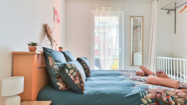 Ein Schlafzimmer mit einem Bett, einem Nachttisch mit Lampe, einem Schminktisch, einem Spiegel und einem Fenster mit Blick auf Gebäude.