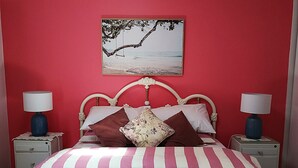 Une chambre avec un mur rouge, un lit avec une literie à rayures, deux tables de chevet avec des lampes et un tableau sur le thème de la plage au-dessus du lit.