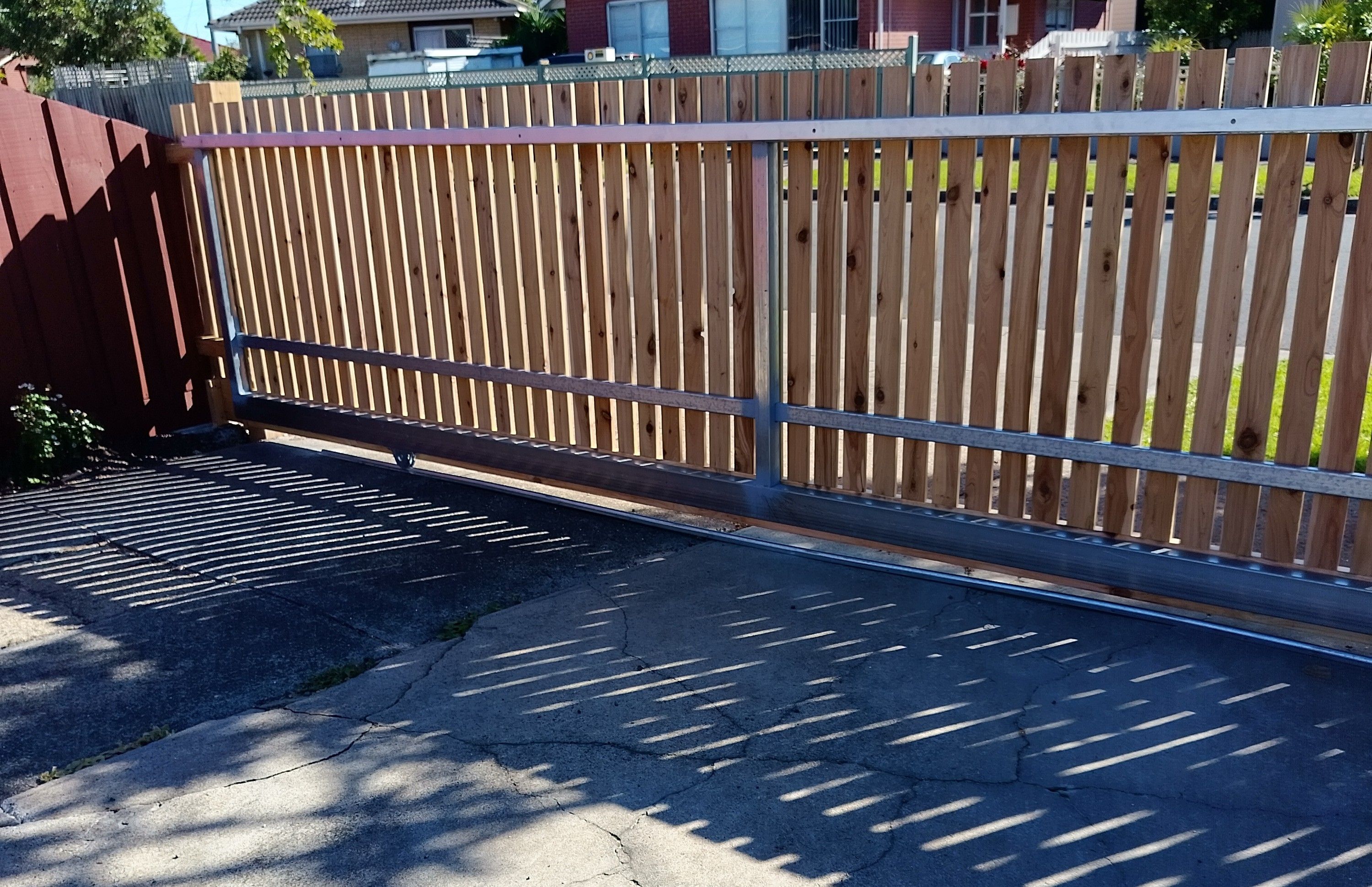 A wooden fence with a metal gate, a paved driveway, and a residential area in the background.