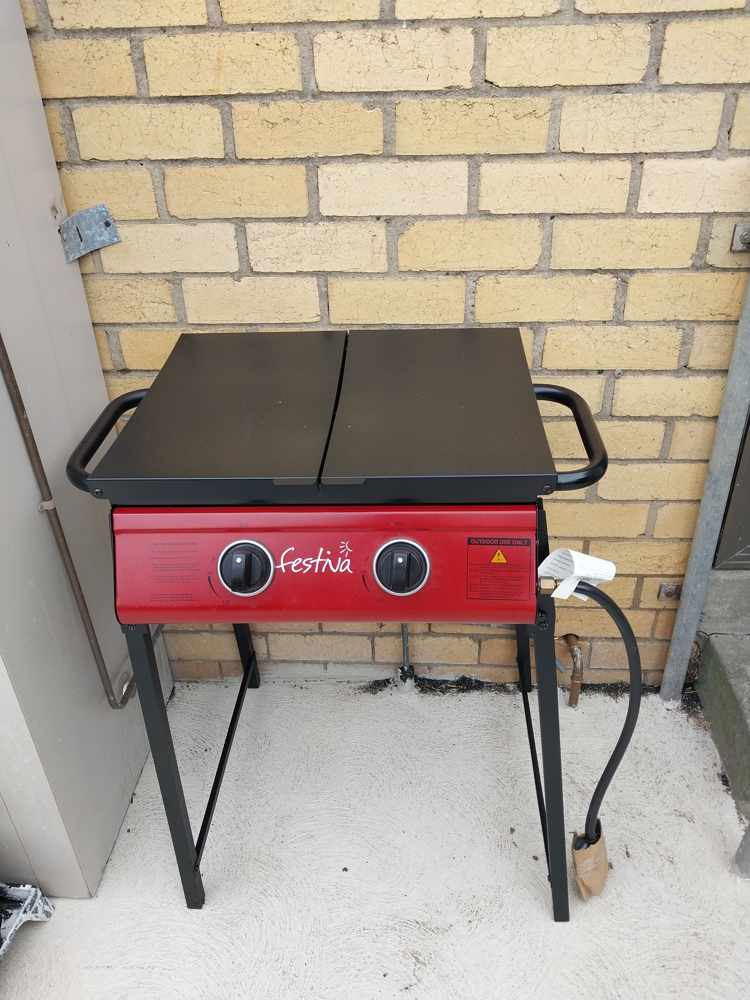 Una parrilla portátil para exteriores con cuerpo rojo y parte superior negra, etiquetada 'festina', frente a un muro de ladrillo.