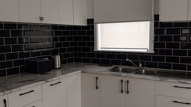 Een moderne keuken met witte kasten, een zwart betegelde spatwand en een raam met jaloezieën.