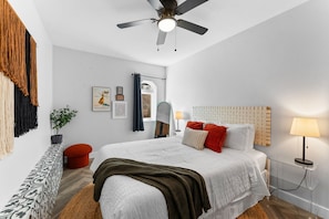 Quarto moderno com cama, ventilador de teto, mesa de cabeceira com abajur, planta em vaso e parede com quadros.