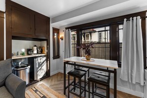 Uma cozinha compacta com armários de madeira escura, bancada de mármore branco e uma janela com cortinas brancas.