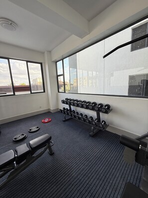 A gym with weightlifting equipment, a bench, and a large window with a view of buildings. - Modern Airport Studio Near JKIA, SGR, Nairobi Expy (Syokimau)