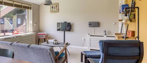 A modern living room with a sofa, a coffee table, and a television. There is a window with blinds, a lamp, and a shelf with books and decorative items.