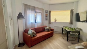 A compact living space with a red sofa, a dining table with chairs, a floor lamp, and a window with curtains.