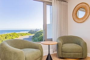 Une pièce comprenant deux fauteuils verts, une petite table ronde, une grande fenêtre donnant sur la plage et l’océan, et un miroir rond accroché au mur.